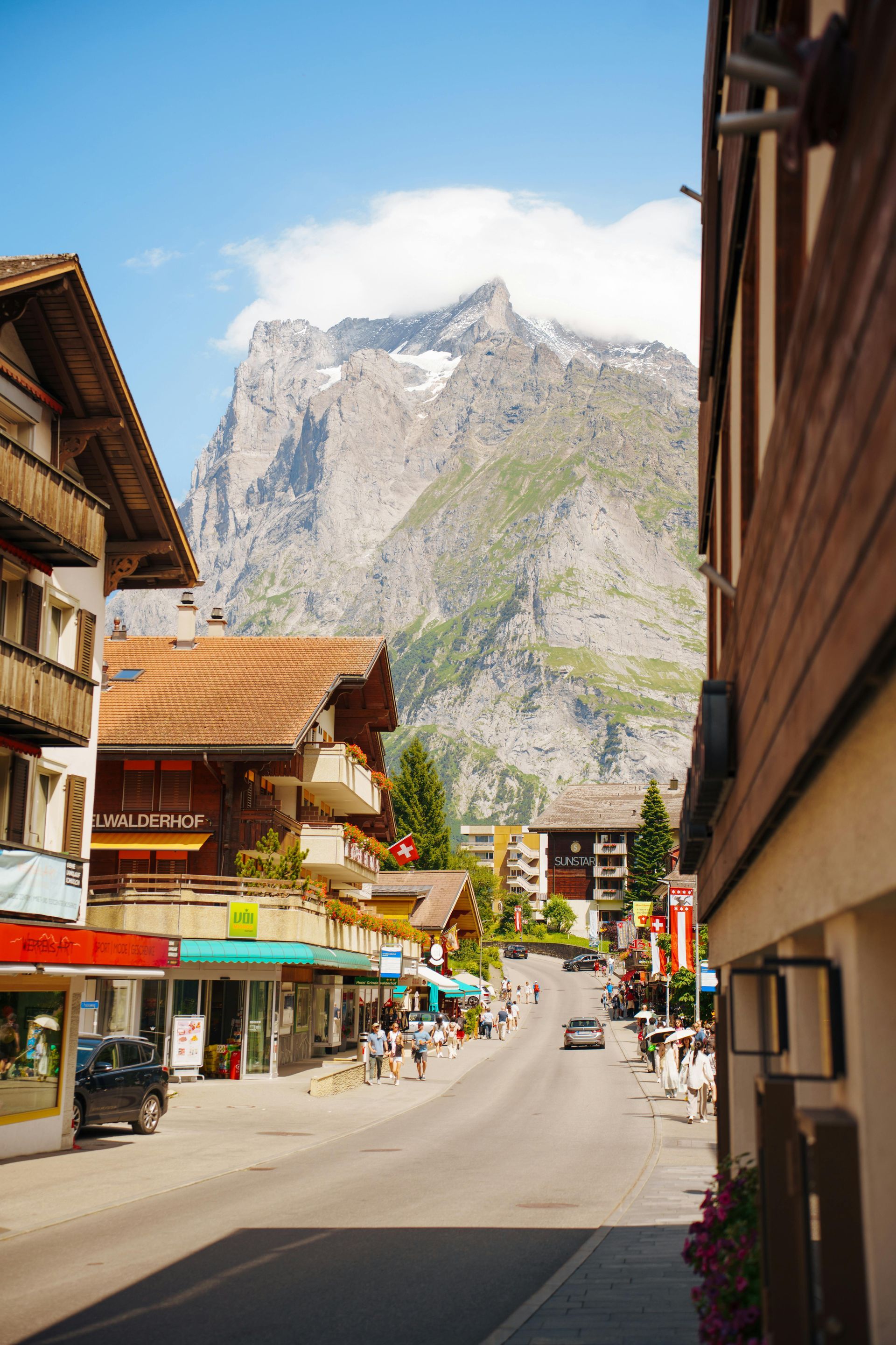 Mountain village street with wooden buildings and shops, leading toward a rocky alpine peak under a blue sky.