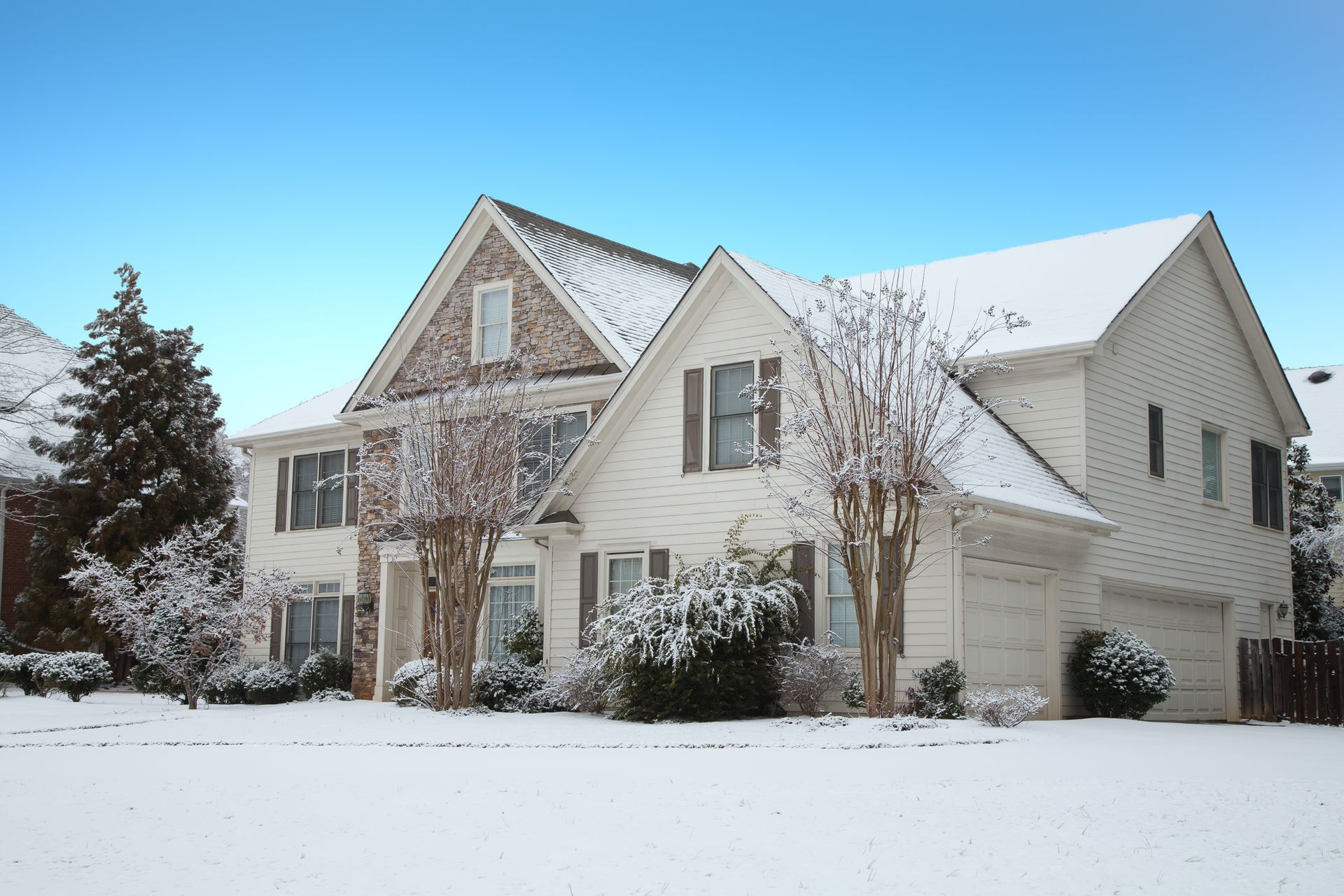 Maison blanche recouverte de neige avec des accents de briques et des arbres sans feuilles sous un ciel bleu vif.