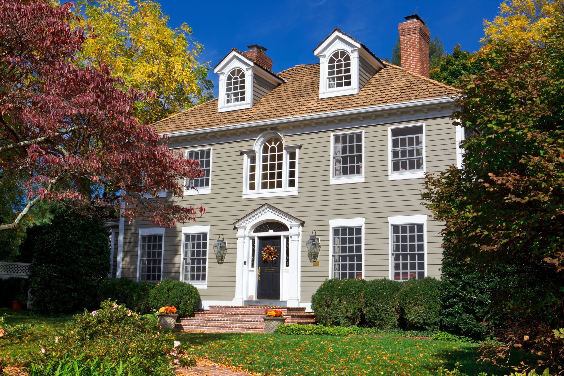 Une maison de style colonial, de deux étages, gris clair, avec des boiseries blanches, une porte d'entrée noire et deux lucarnes, sous un ciel bleu.
