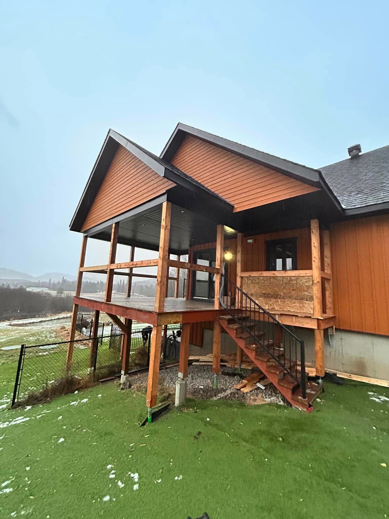 Une véranda en bois avec un toit en bardeaux et un escalier, attenante à une maison donnant sur une cour herbeuse légèrement enneigée.