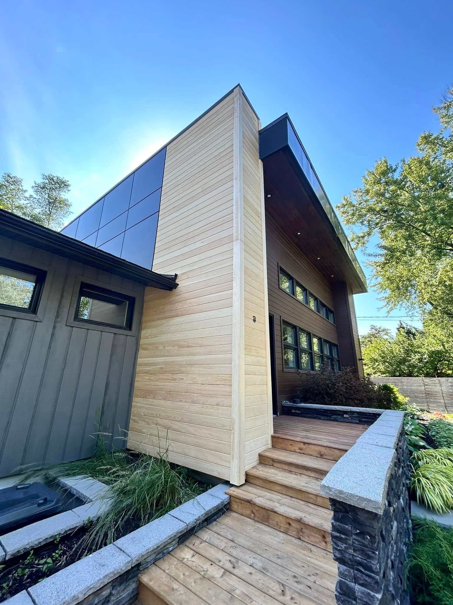 L'entrée d'une maison moderne, avec une façade en bois clair, un bardage métallique foncé, des marches et un mur de soutènement en pierre, sous un ciel bleu.