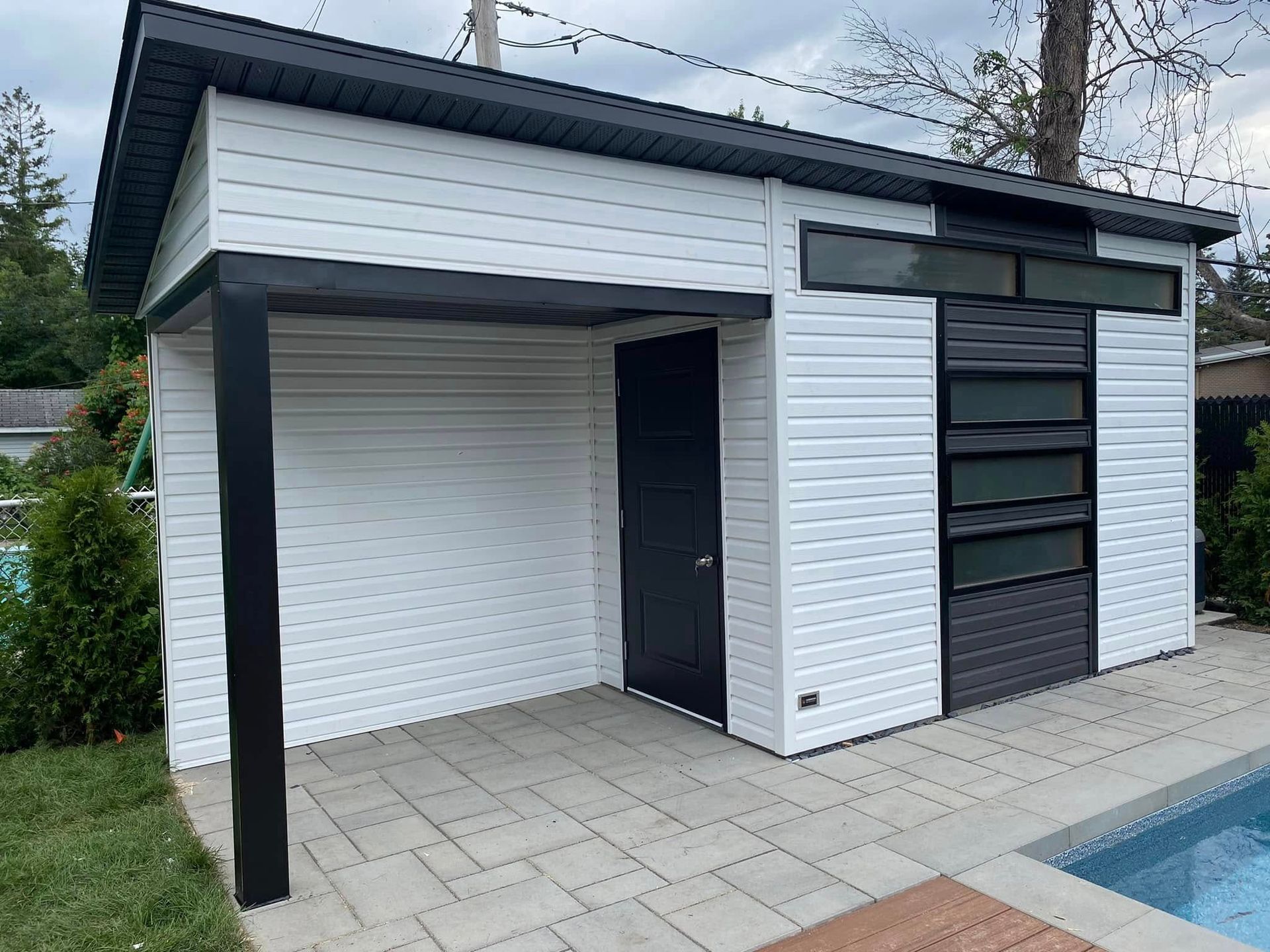 Un abri de jardin blanc moderne avec une porte noire, une poutre de soutien noire et une rangée de fenêtres horizontales, situé à côté d'une piscine.
