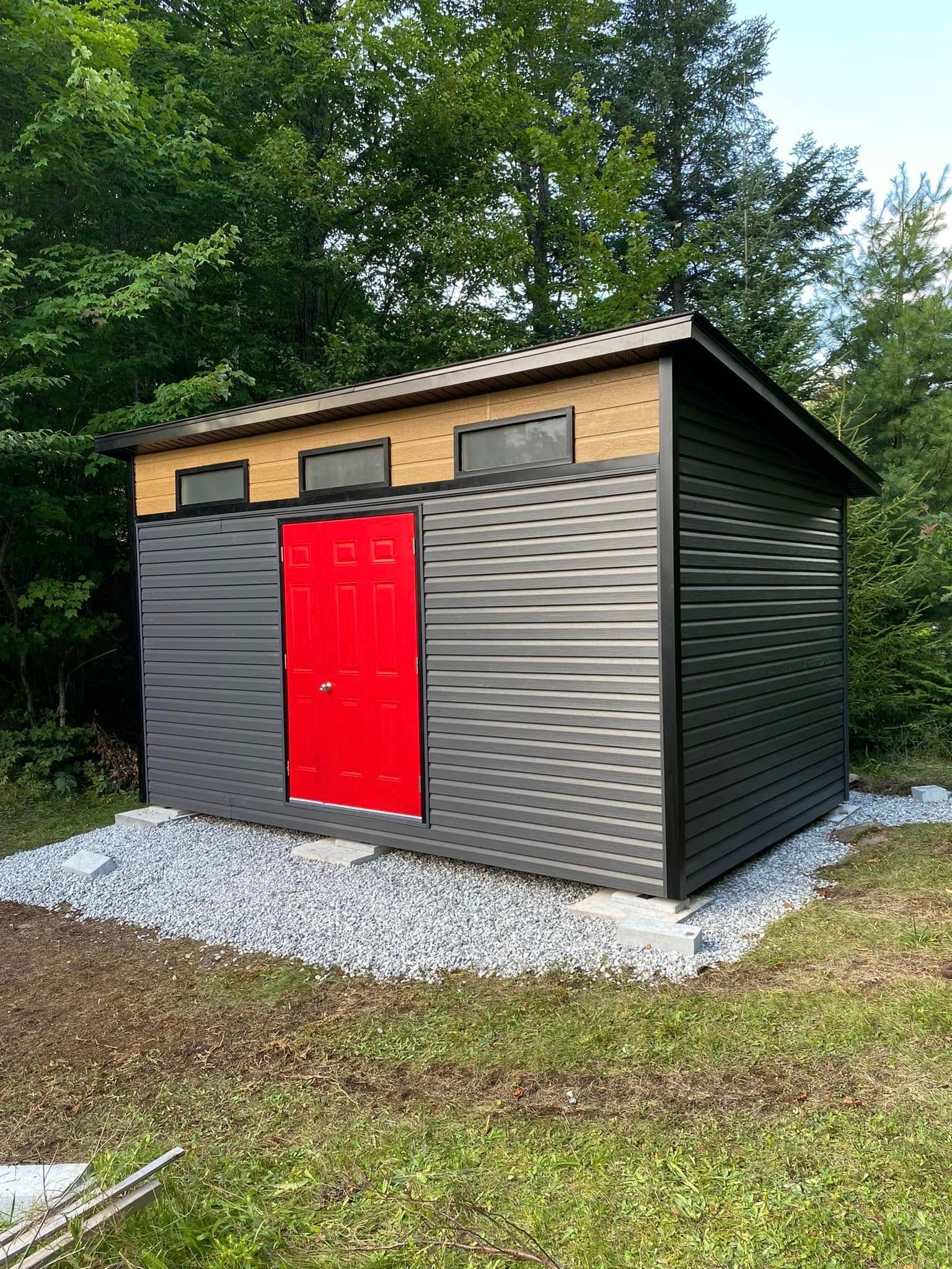 Abri de jardin moderne avec bardage en tôle ondulée noire, une porte rouge vif et trois fenêtres de toit, installé sur une dalle de gravier.