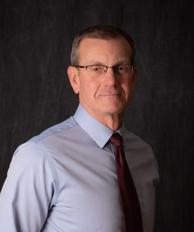 A man wearing glasses and a tie is standing in front of a black background.