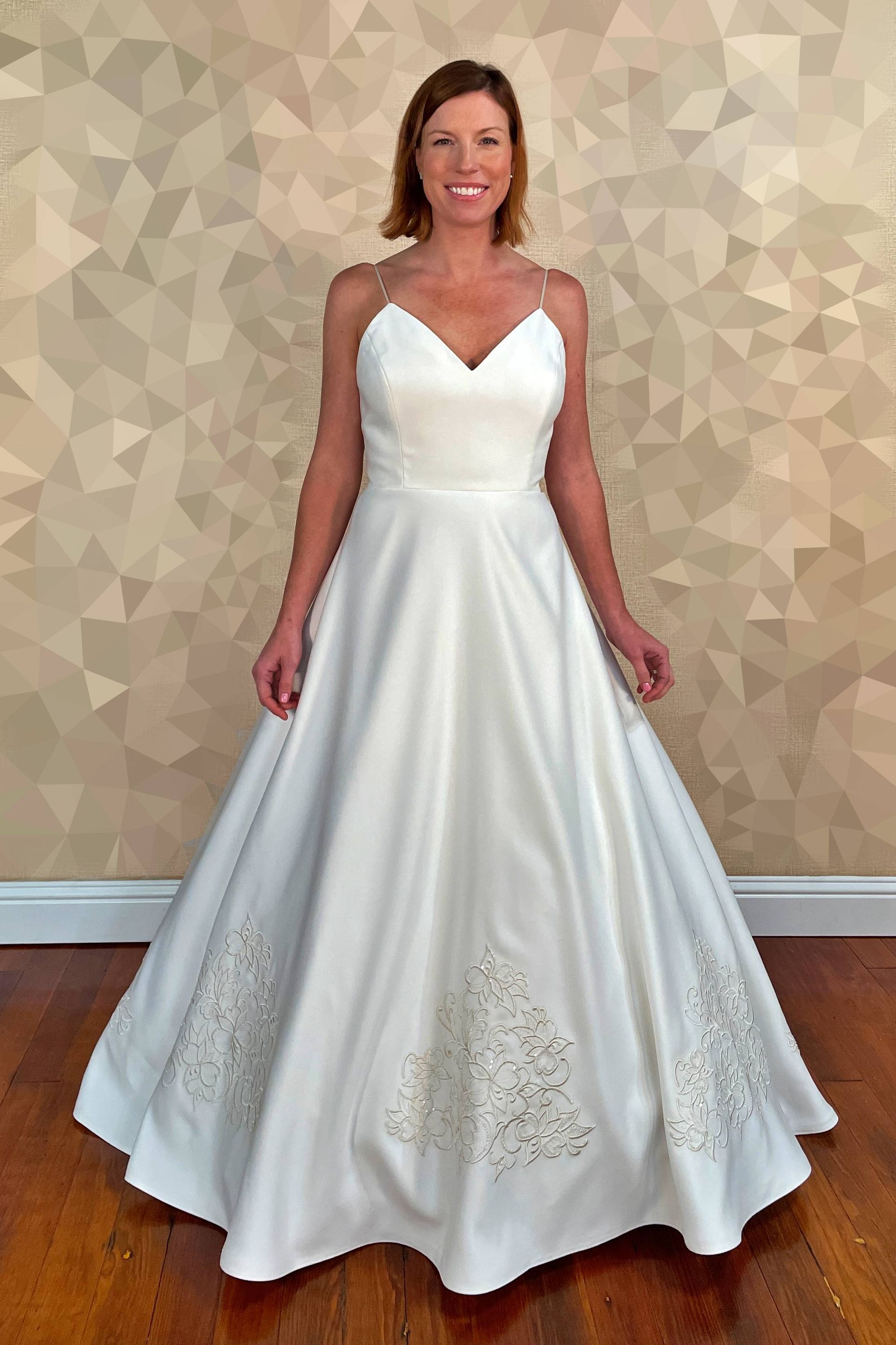 Woman in a white wedding gown smiles, standing in front of a textured wall and wooden floor.