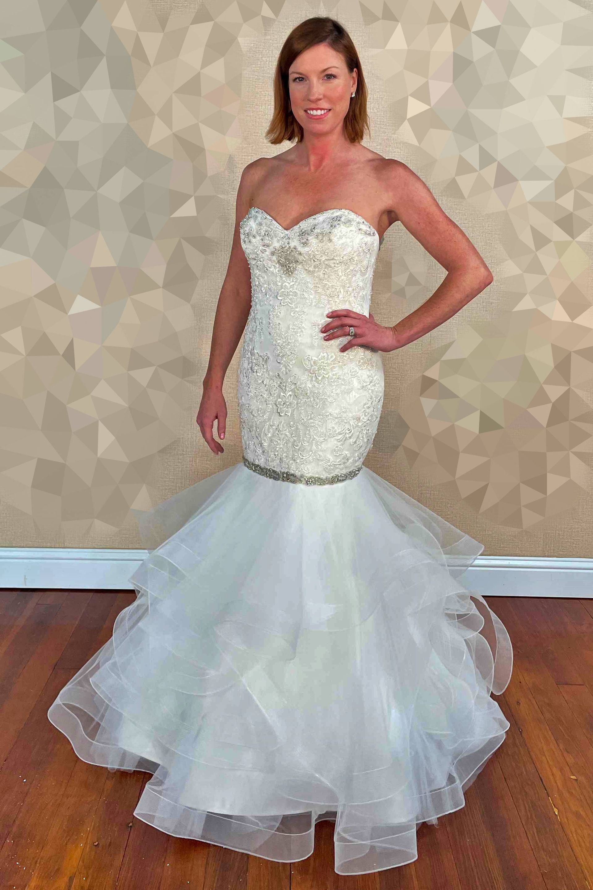 Woman in a strapless white beaded wedding dress with a tiered, ruffled skirt, standing indoors, smiling.
