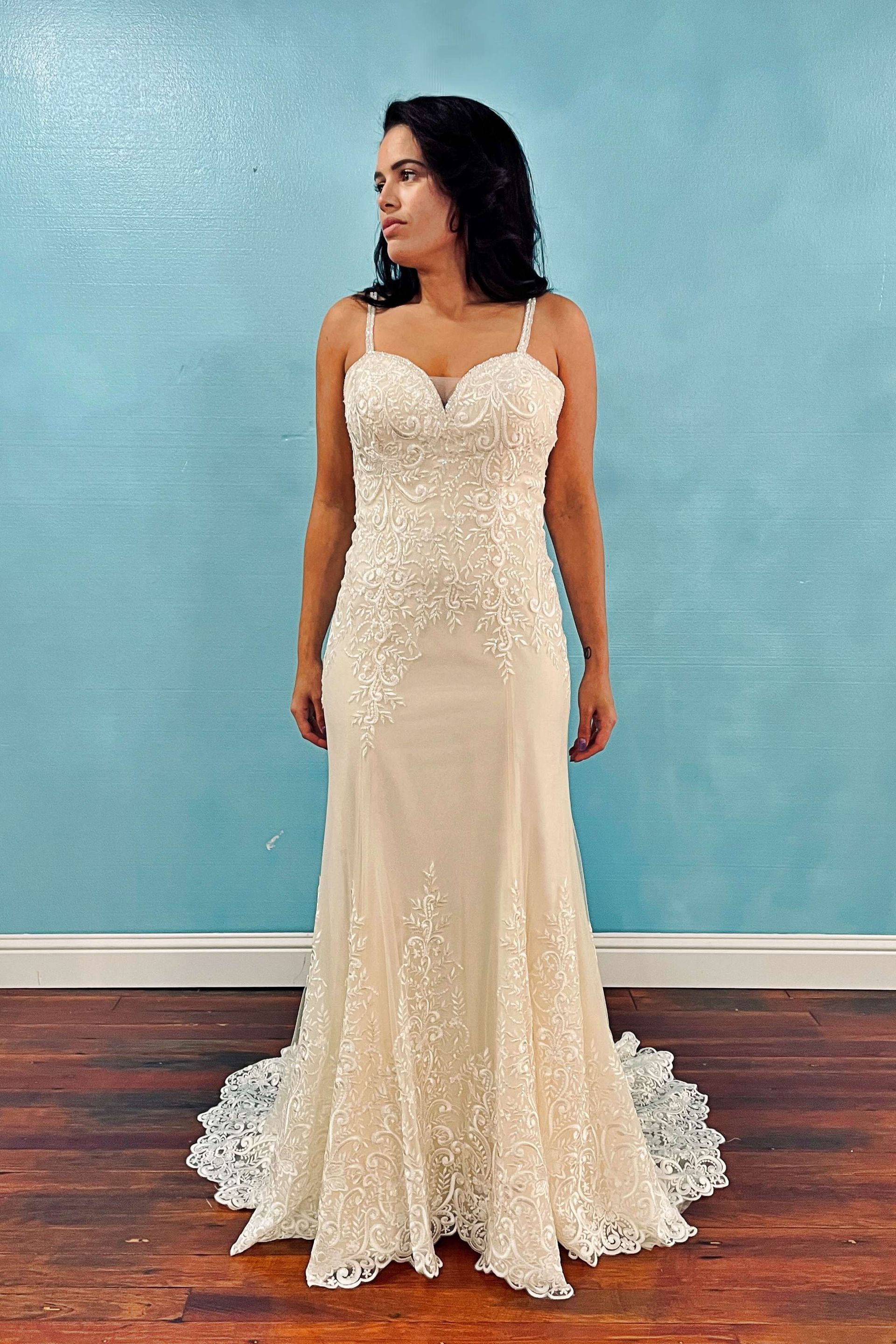 Woman in a wedding dress with beaded bodice, spaghetti straps, and mermaid skirt, standing in front of a blue wall.