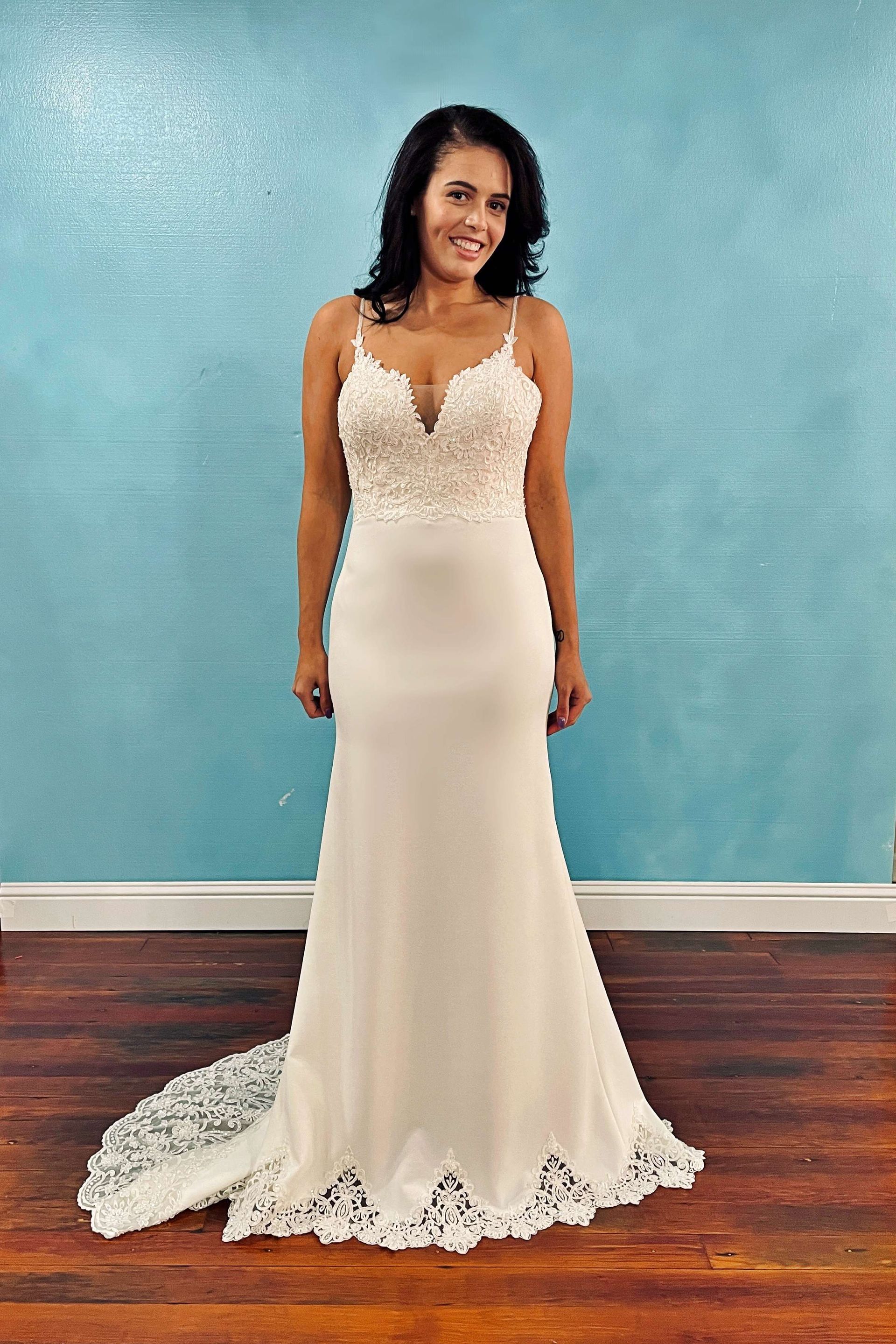 Woman in a white wedding dress with lace bodice, smiling, standing in front of a blue wall.
