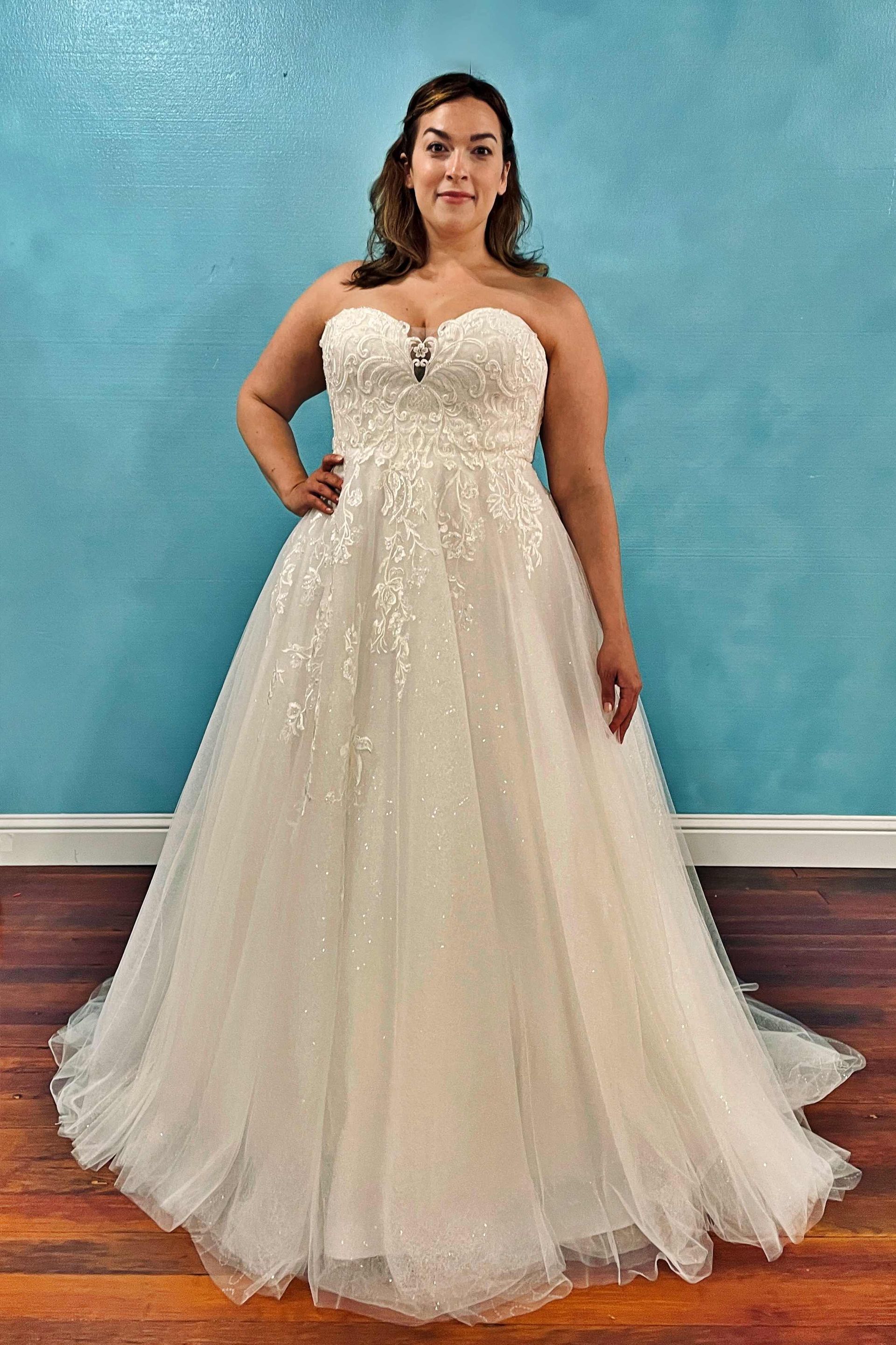 Woman in strapless wedding dress with embellishments, posing in front of a blue wall.