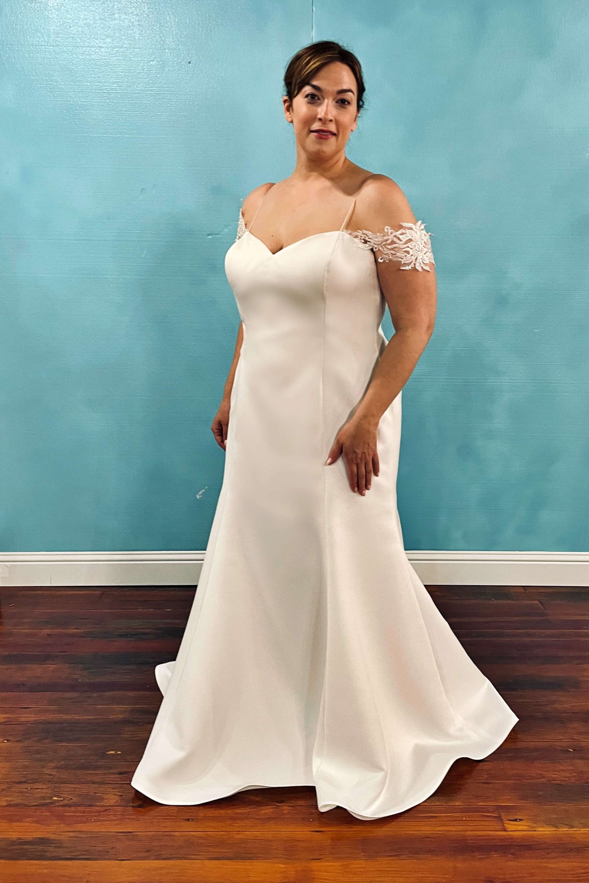 Woman in a white off-shoulder wedding gown poses against a blue wall and wood floor.
