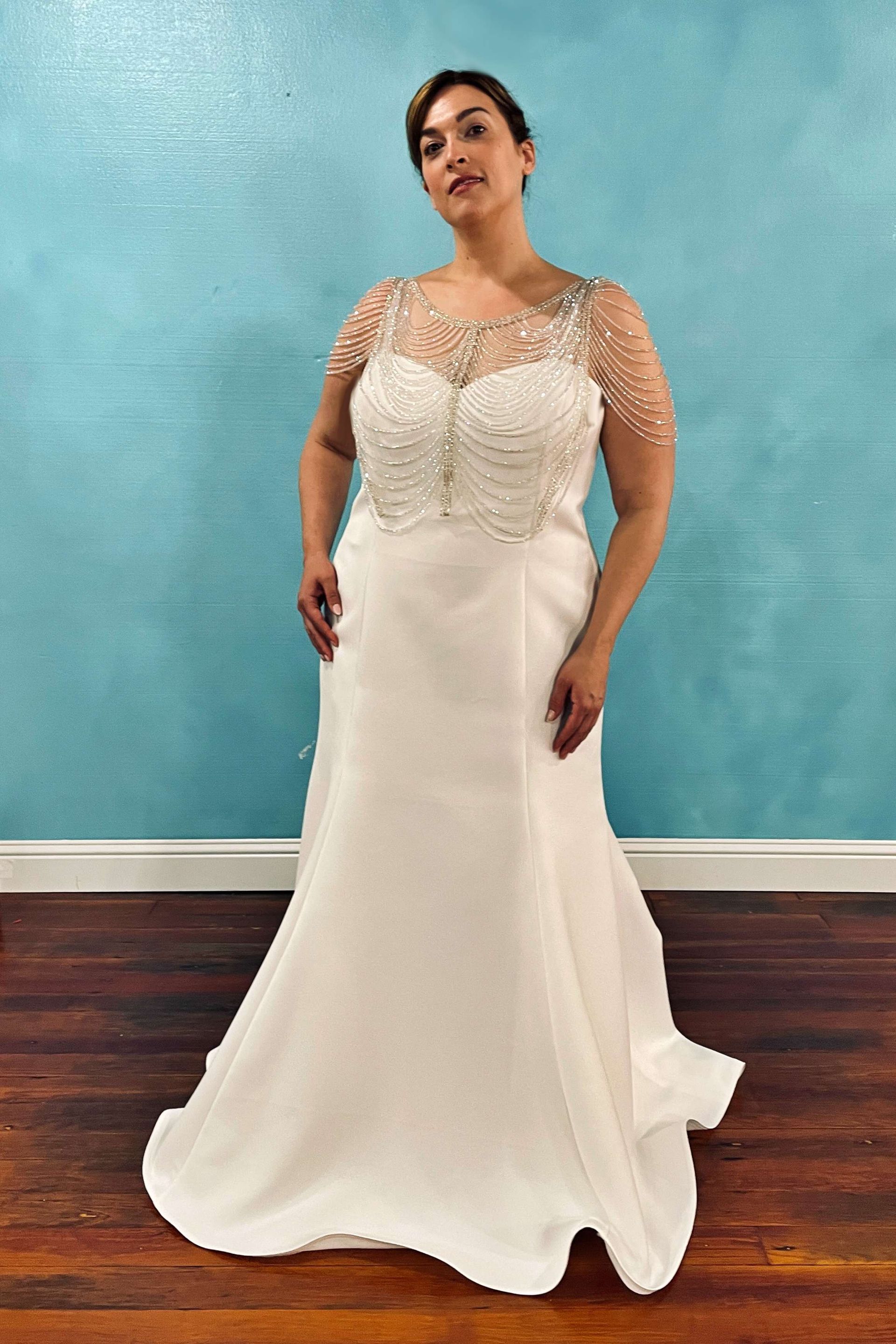 Woman in white wedding dress with beaded bodice, posing against blue wall.