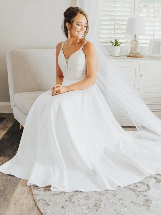 Woman in white strapless wedding dress, smiling, arms outstretched, against a bright white backdrop.
