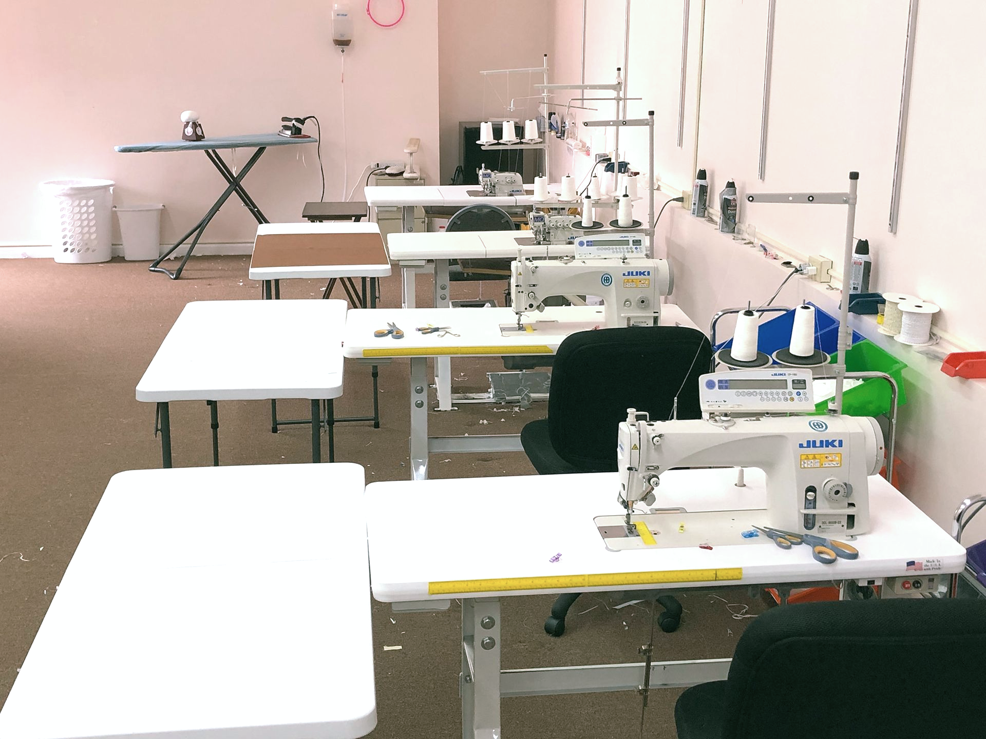 Sewing studio with white tables and machines, set against pale walls, and brown flooring.