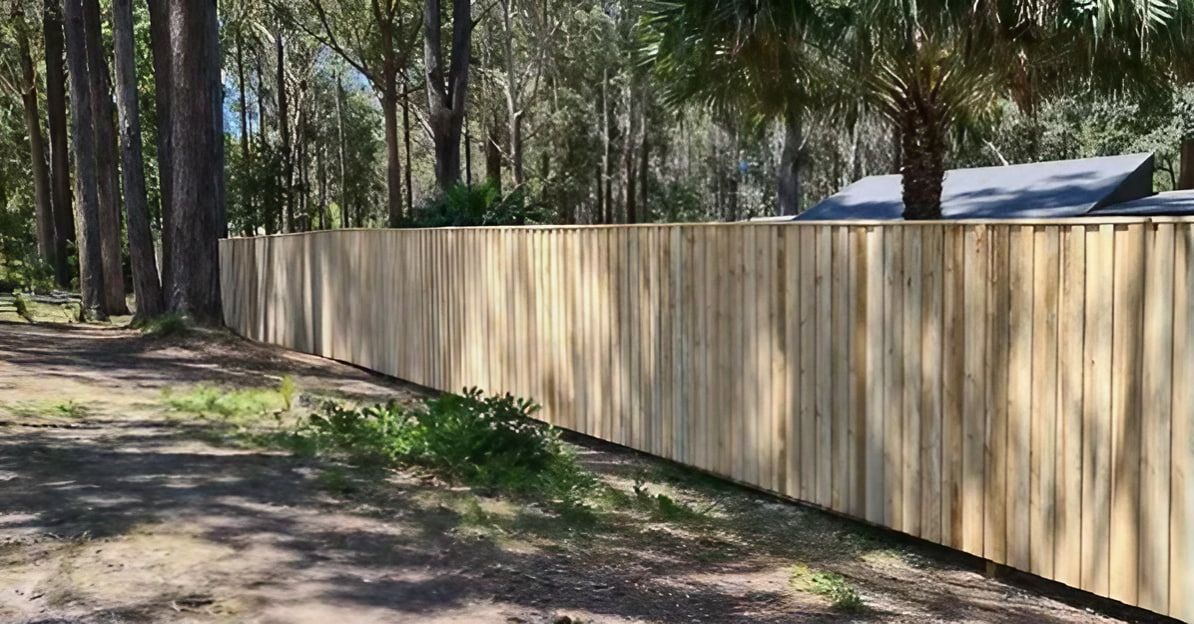 A Wooden Fence Surrounds a Dirt Road in The Middle of A Forest — D & B Fencing in Taree, NSW