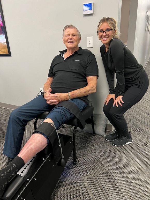 Two people in a hallway, one seated with a leg brace and smiling, the other kneeling beside them.