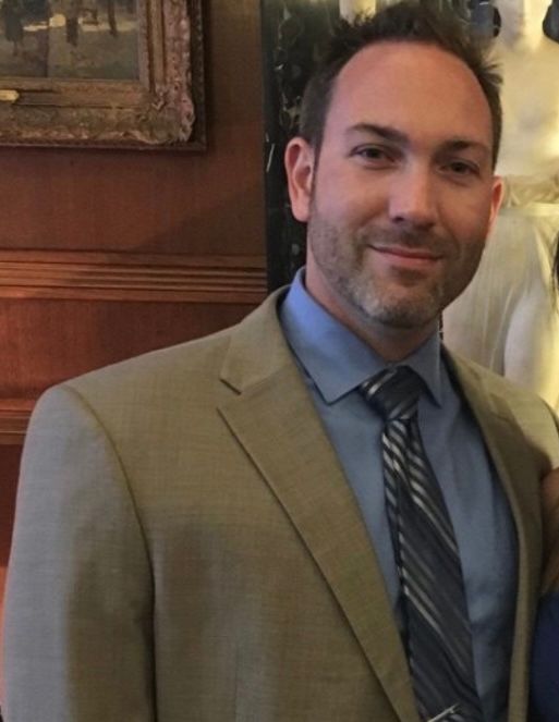 Man in tan suit and blue shirt smiling indoors, with wood-paneled wall behind him