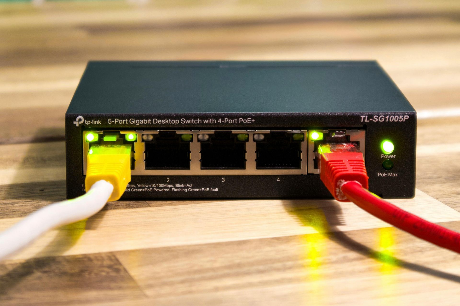 Ethernet switch with white and red network cables plugged in, green status lights on a wooden desk.