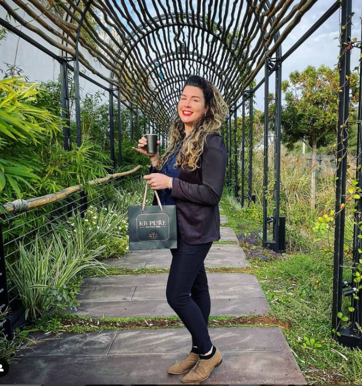 Woman standing on a garden path under an arched trellis, holding a small green handbag and smiling.