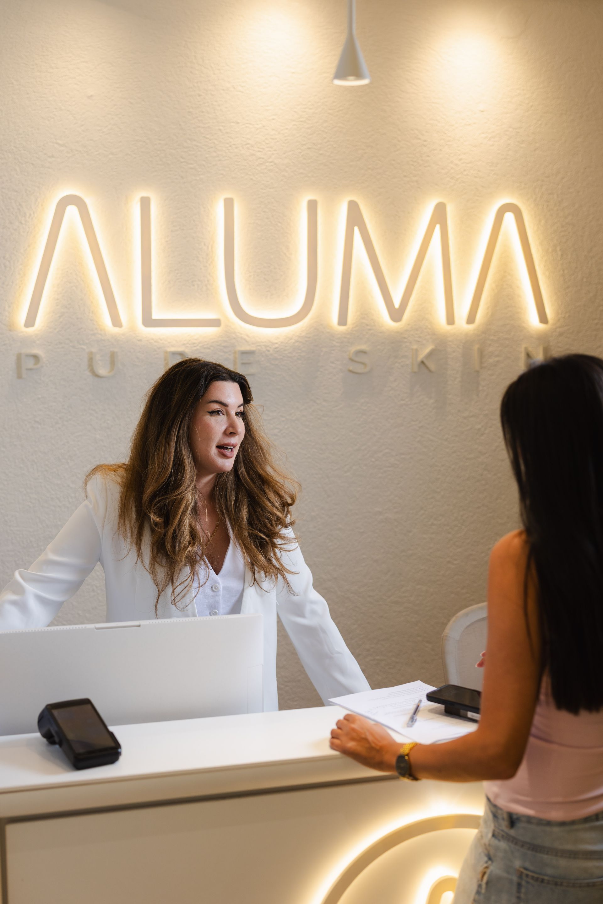 Reception desk with illuminated ALUMA sign and two women talking in a lobby