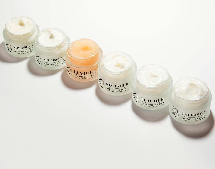 Five small cosmetic jars in white and peach arranged diagonally on a light background.