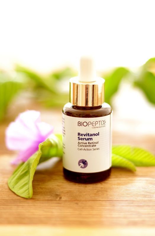 Bottle of BIOPEPTIDE Serum with dropper on a wooden table, surrounded by soft green leaves and a pink flower