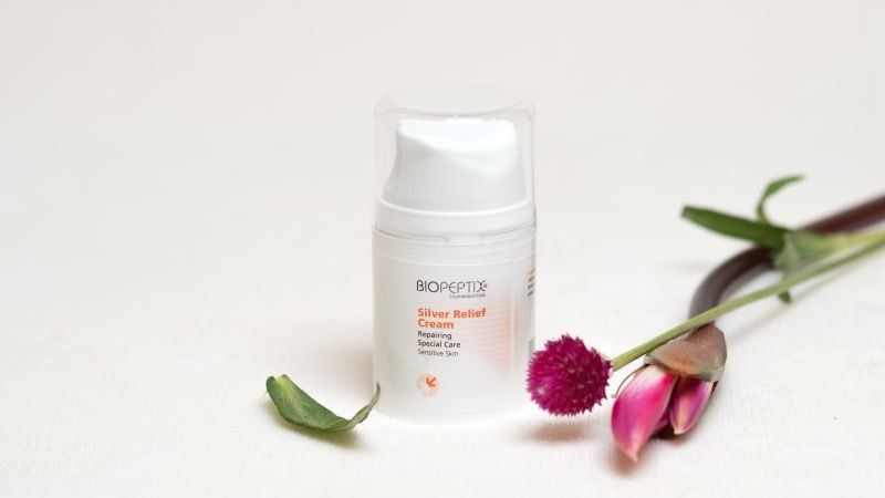 White cosmetic bottle beside pink flowers and green leaves on a light background