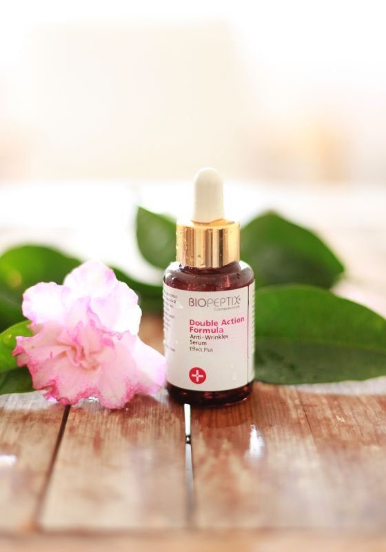 Brown dropper bottle with pink label beside a pink flower and green leaves on a wooden surface