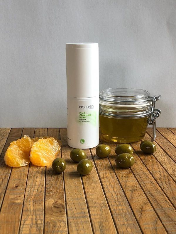 White cosmetic bottle beside jar of oil, orange slices, and green olives on a wooden table.