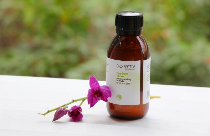 Brown bottle with a white label beside a purple flower on a white surface outdoors