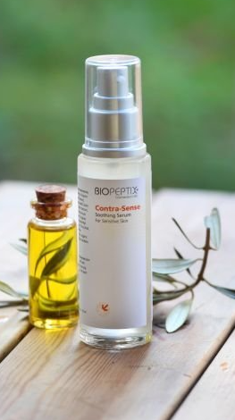 Silver cosmetic spray bottle beside amber oil bottle on a wooden table with greenery in the background