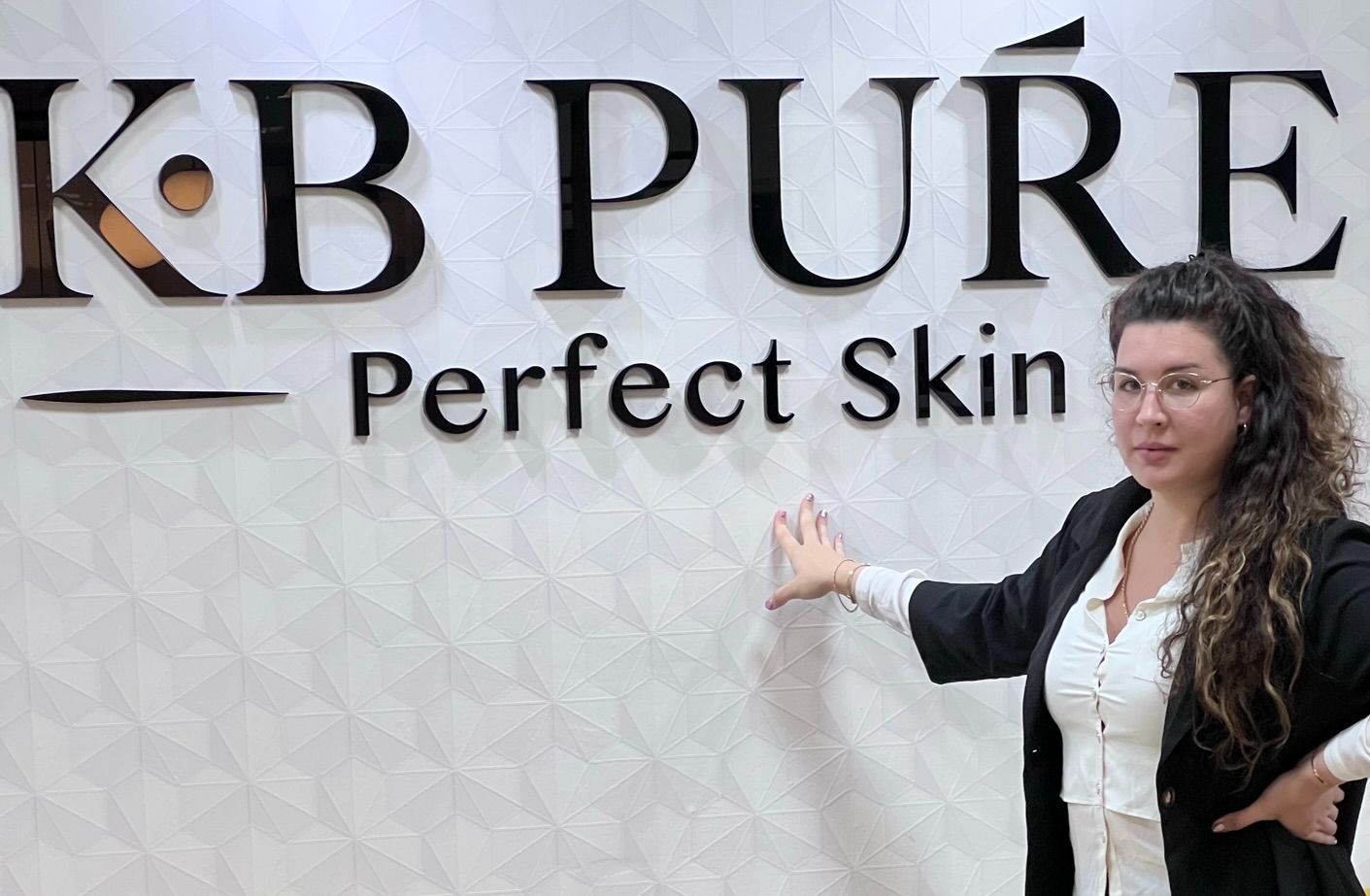 Woman standing beside “KB PURE Perfect Skin” sign on a white textured wall