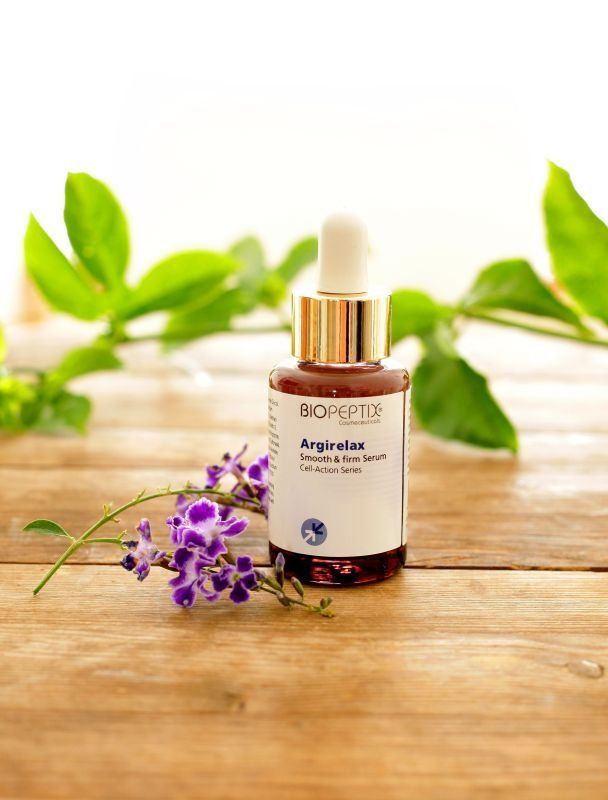 Bio-ethereal argan oil dropper bottle on a wooden table with purple flowers and green leaves