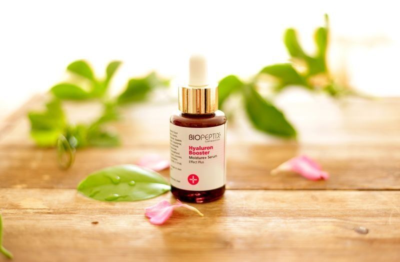 Brown essential oil dropper bottle on a wooden table with green leaves and pink petals in soft light