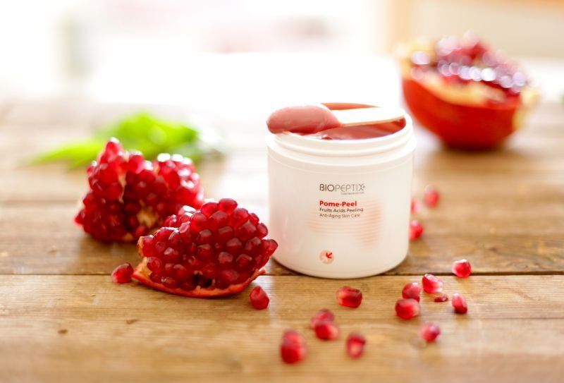 White cosmetic jar with raspberries on a wooden table, with scattered berries and a blurred bowl in the background