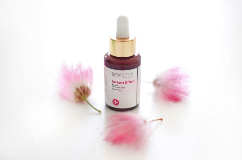 Pink facial serum bottle with gold dropper surrounded by pink flowers on a white background