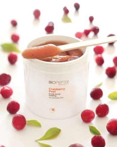 White jar of cranberry lip balm with a spoon, surrounded by cranberries and green leaves on a white background