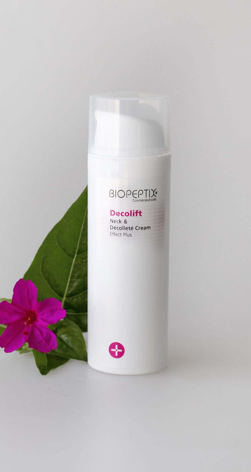 White cosmetic bottle with pump beside a pink flower and green leaf on a light background