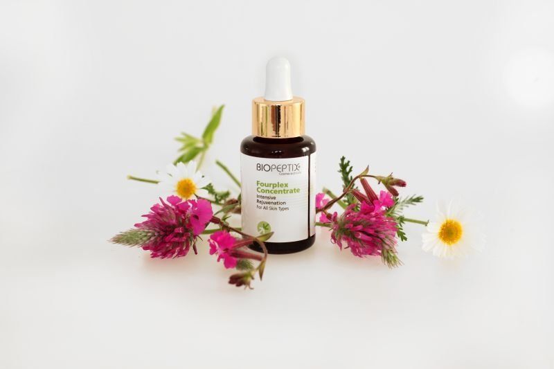 Brown skincare dropper bottle with gold cap, surrounded by pink flowers on a white background