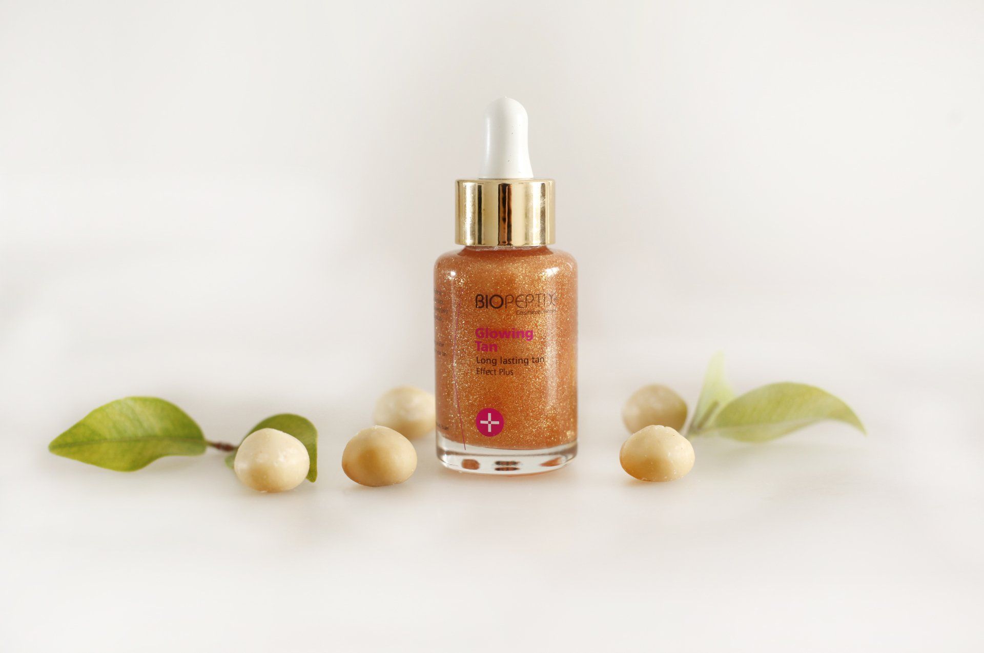 Amber serum bottle with dropper, flanked by green leaves and cream-colored stones on a white background