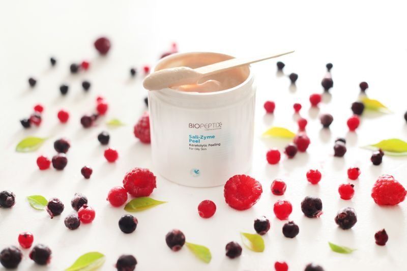 White skincare jar with peach cream, surrounded by scattered colorful berries on a white surface