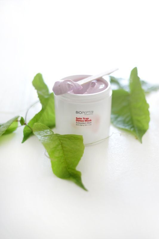 White skincare jar with lavender lid beside green leaves on a bright white background