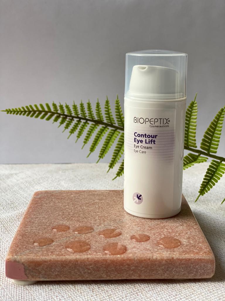 White skincare bottle with clear cap on a pink stone block, beside a fern leaf on a gray background.