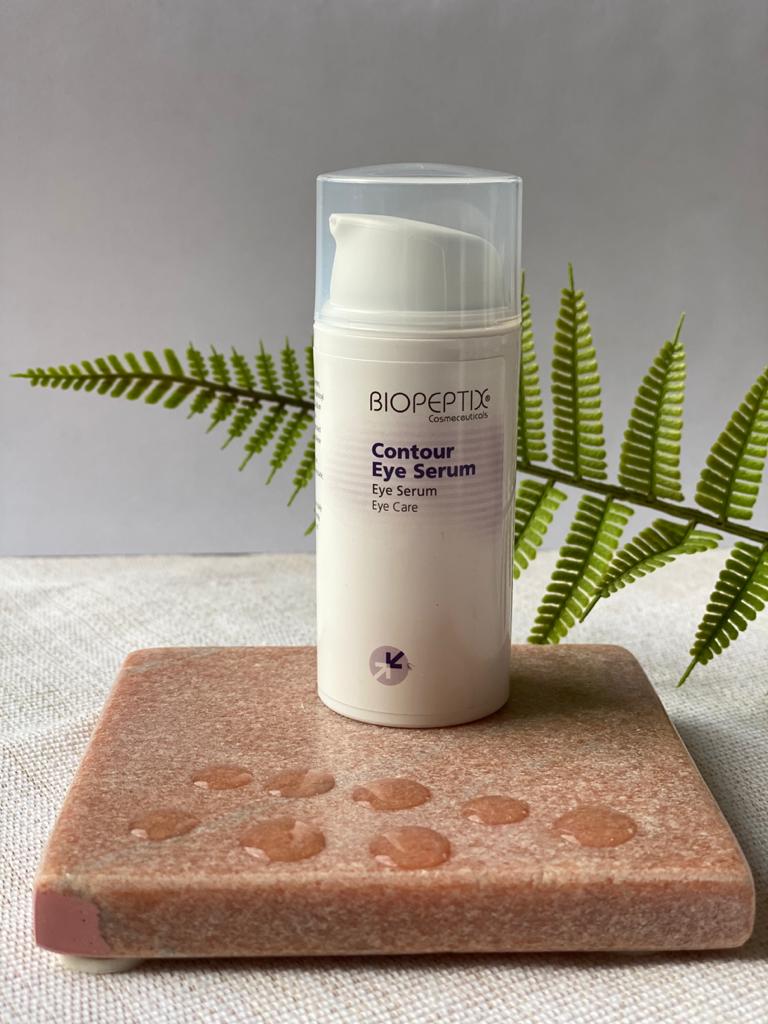 White skincare serum bottle with clear cap on a pink stone slab, framed by fern leaves.