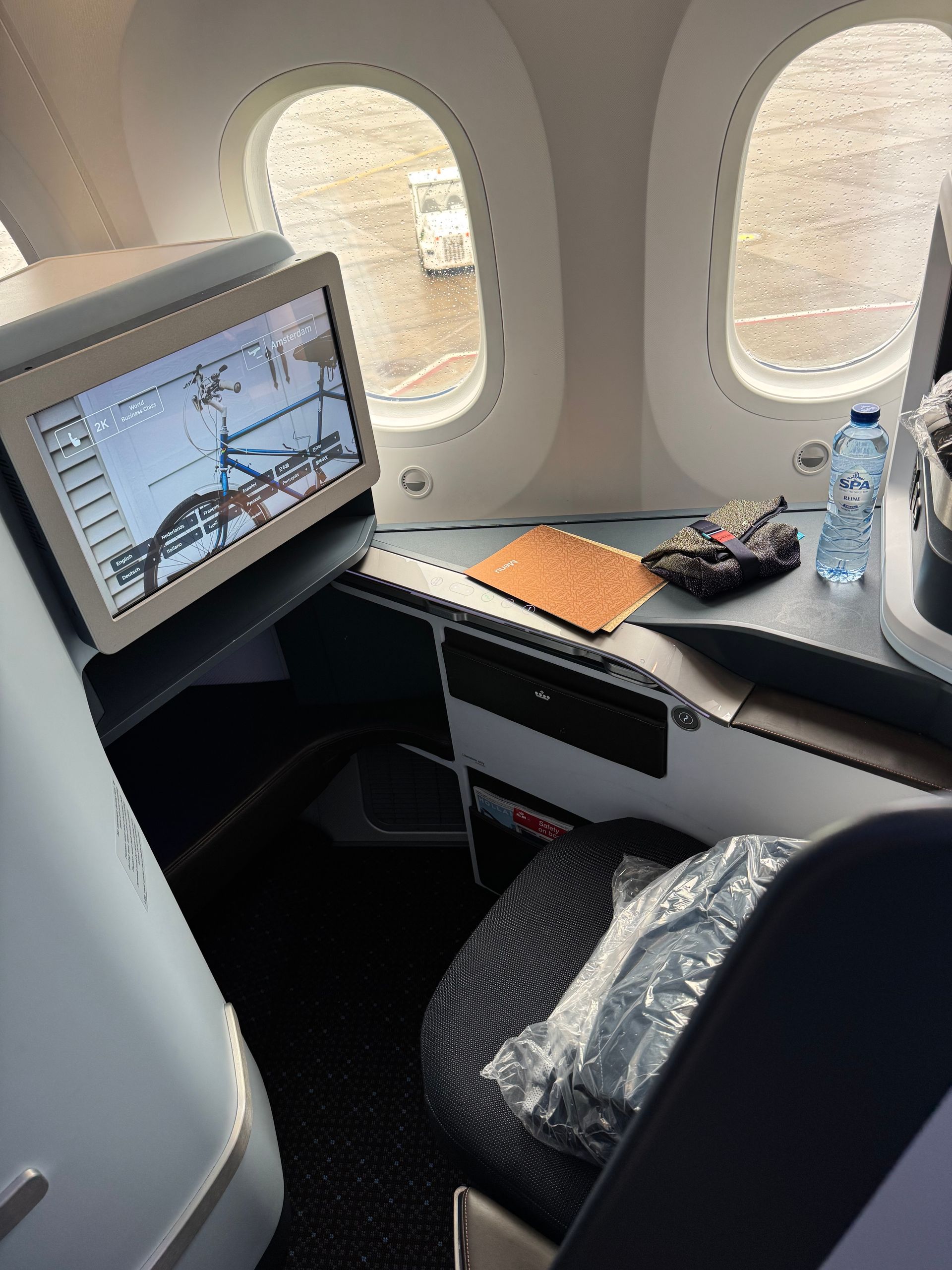Airplane business-class seat with in-flight entertainment screen, tray table, notebook, headphones, and water bottle
