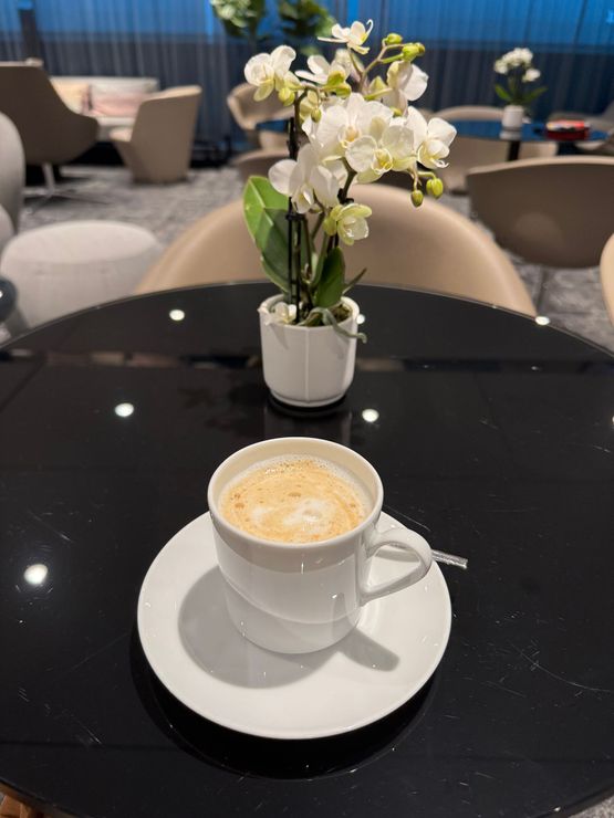 White coffee cup on saucer with latte art on a glossy black table, with white flowers in a vase behind it