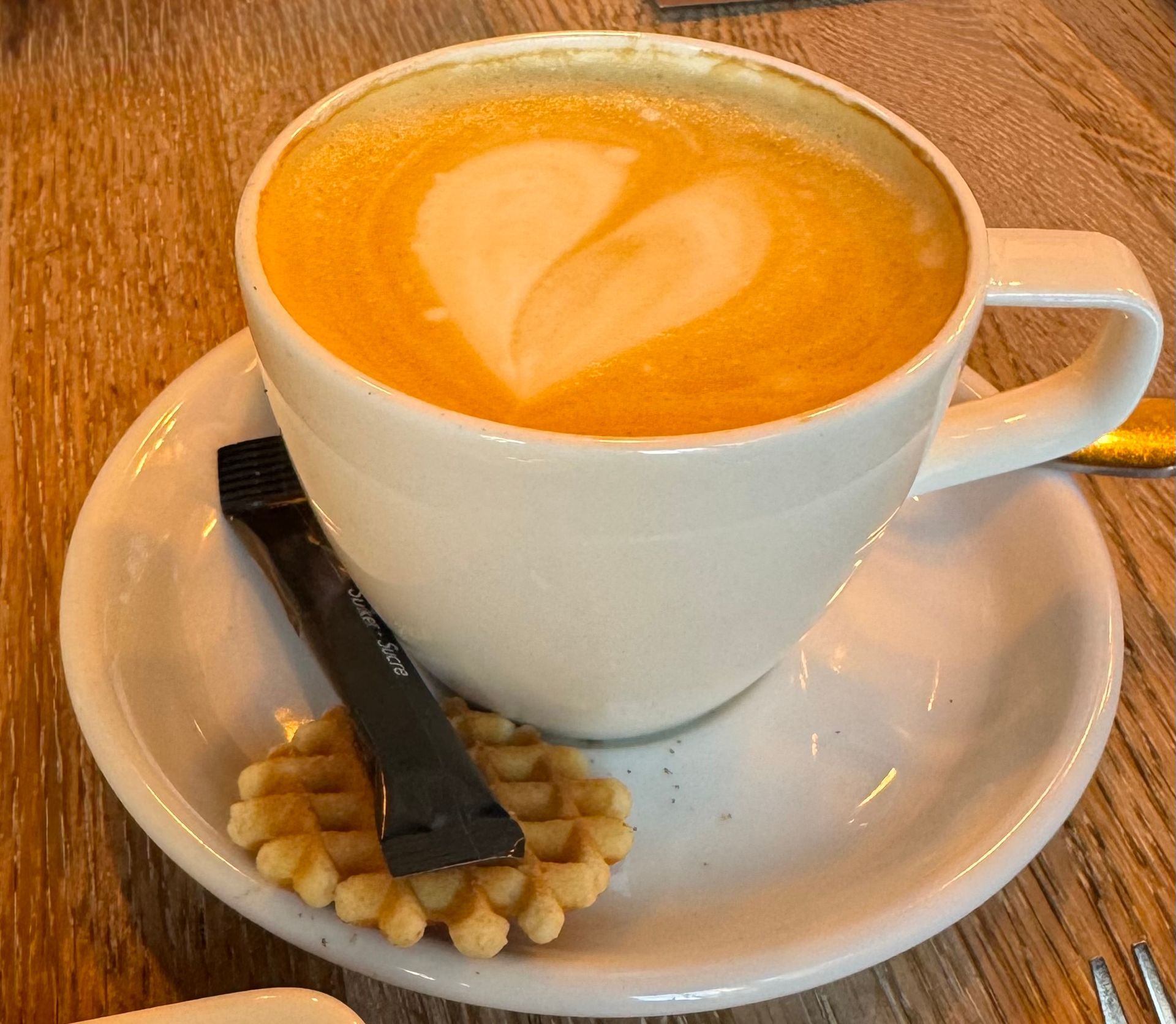 White coffee cup with latte art on a saucer, beside a waffle and black spoon on a wooden table