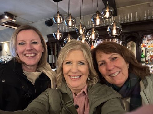 Three smiling friends posing for a selfie in a cozy bar with hanging lights