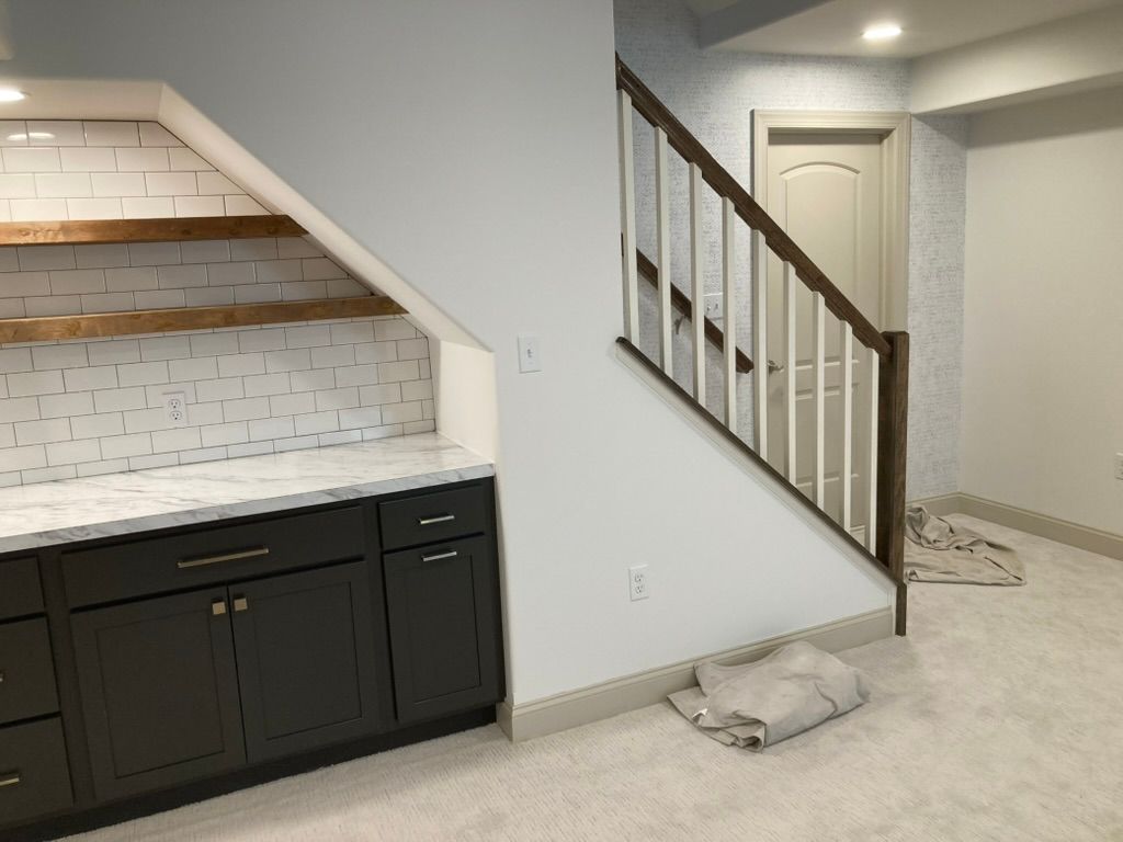 Built-in bar area under stairs, with cabinets, marble countertop, white brick tile, and a wooden stairwell.