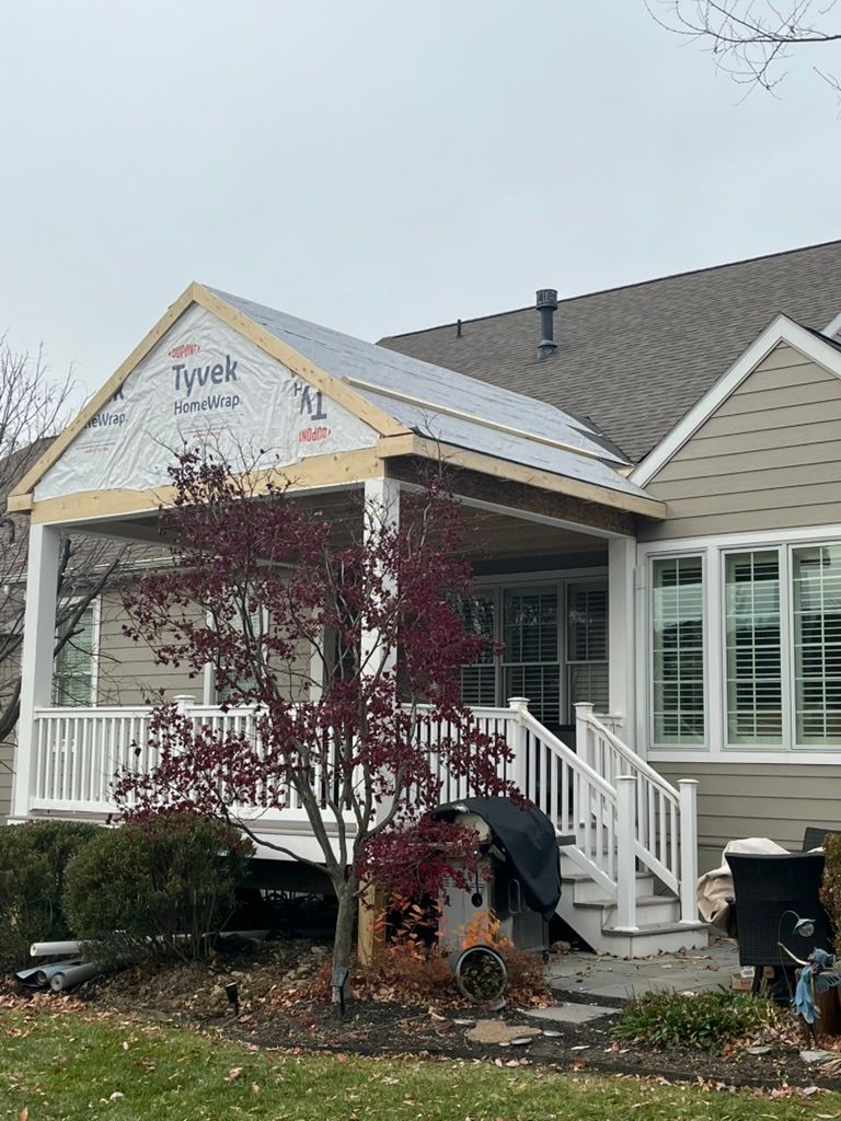 A house with a partially constructed covered porch. Exterior features beige siding, white trim, and a small tree.