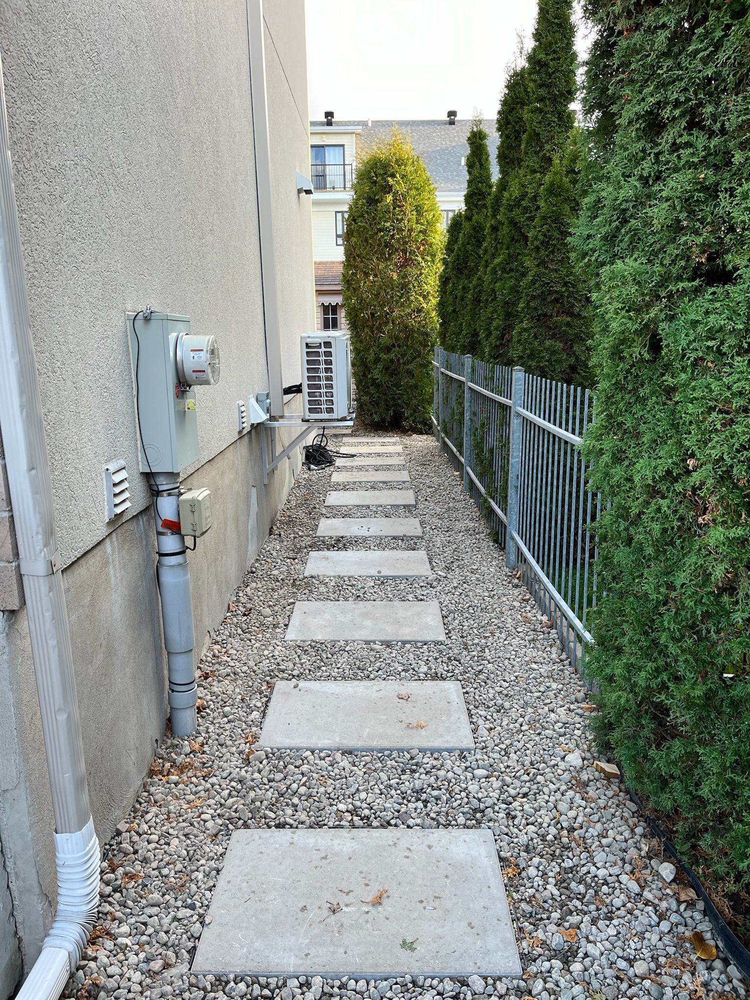 Des dalles de béton permettent de traverser une étroite cour latérale recouverte de gravier, bordée d'une clôture et de grands conifères.